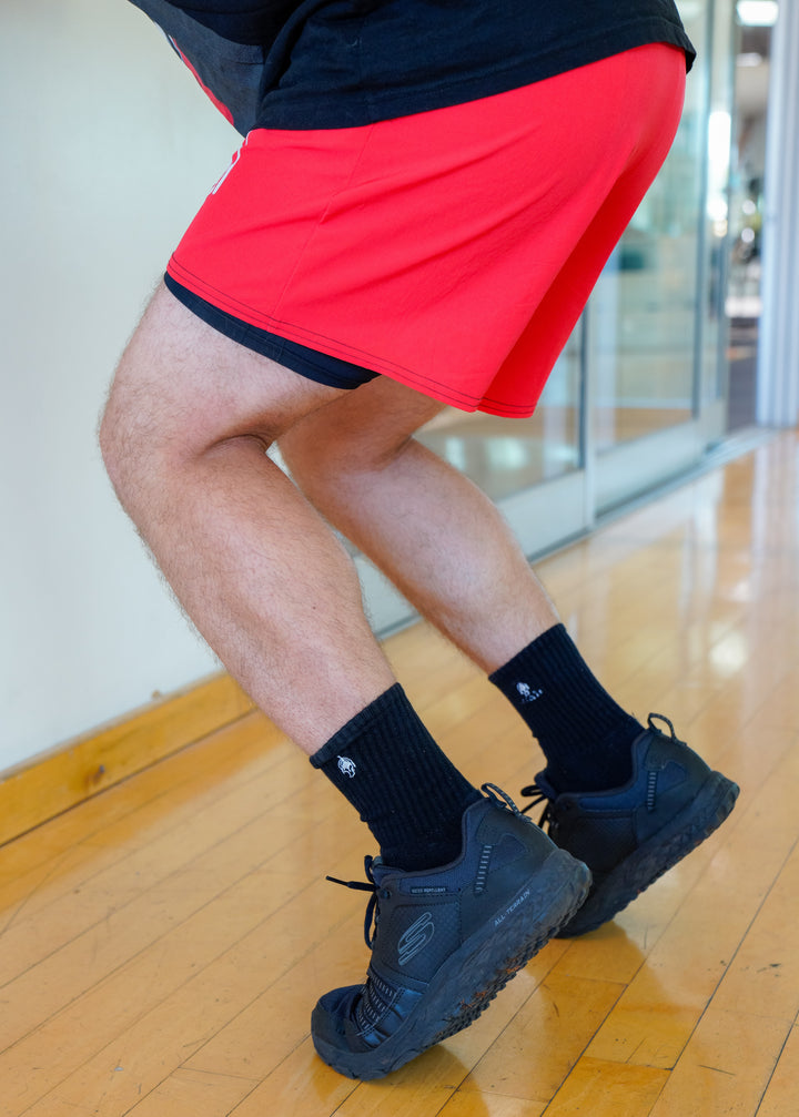 Close-up of Jamin Fitness branded black athletic socks with spartan helmet logo, shown with red workout shorts and black training shoes on wooden gym floor, demonstrating athletic wear quality