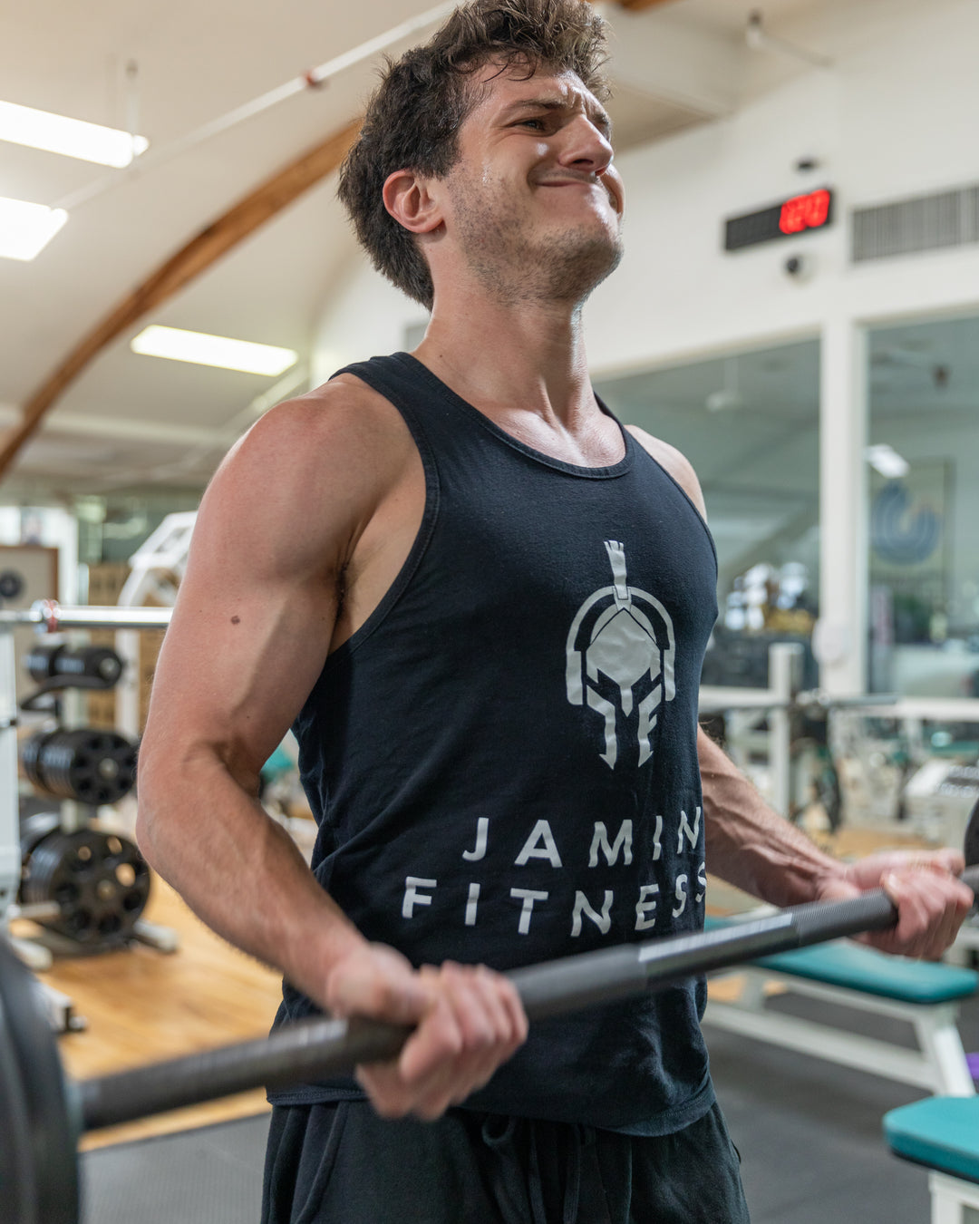 A fitness trainer wearing a black Jamin Fitness tank top. photographed in a gym setting while holding a barbell. The image shows a close-up of the upper body, highlighting the branded apparel and professional gym environment with equipment visible in the background.