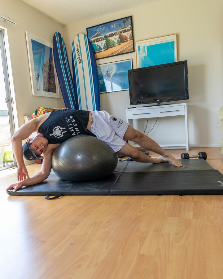 A person demonstrating a flexibility exercise on a black exercise mat, lying sideways over a large grey stability ball. They're wearing a black Jamin Fitness tank top and white shorts. The room has a beach-themed decor with surfboards and coastal artwork, including VW van prints and surfing photos. A TV and white media console are visible in the background