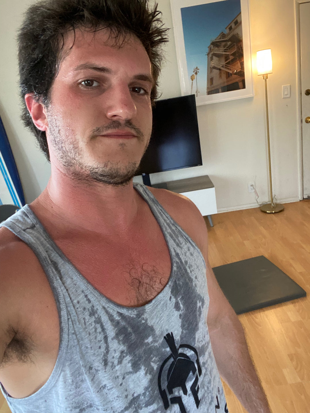 Benjamin Villers wearing gray tie-dye tank top with Jamin Fitness spartan logo, photographed in home gym setting with surfboard art and training mat, showcasing authentic post-workout content